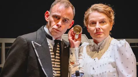 Andrew Billington A man with short hair and wearing a black jacket over a striped waistcoat, white shirt and back tie, stands next to a woman with blonde hair and a white top. Both have confused and surprised expressions on their faces as they both listen to the earpiece of an old fashioned telephone.