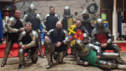 Medieval Combat Wales Group of nine men posing indoors in a training hall, wearing medieval-style metal armour over dark clothing. Some kneel while others stand behind them. One person at the front holds a painted shield with bold red, yellow, and black sections. The room has wooden panelling, a stone wall, padded mats, and hanging equipment such as gloves and a tyre.
