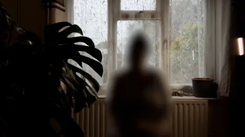Jon Ironmonger A blurred image of a person sitting in a darkened room. Behind them is a window and a radiator. They are in silhouette and their features have been removed to protect their identity. 