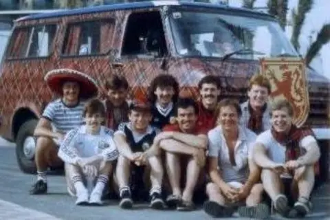 Jim Stewart A group of Scotland fans sitting in front of a campervan which has been covered in tartan wallpaper.