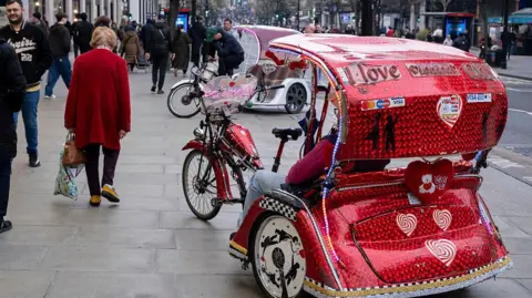 A red rickshaw decorated with hearts, lights, and “I Love London City” text is parked on a busy pavement as pedestrians walk past on a central London street.