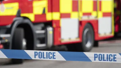 Blue and white police tape in front of a red and yellow fire engine. 