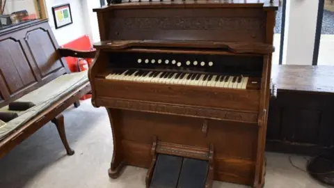 A room which has a church organ that is plated with dark wood. The organ is small and has intricate decorative carvings on it. There are other wooden furniture pieces inside the room which is an auction house. 
