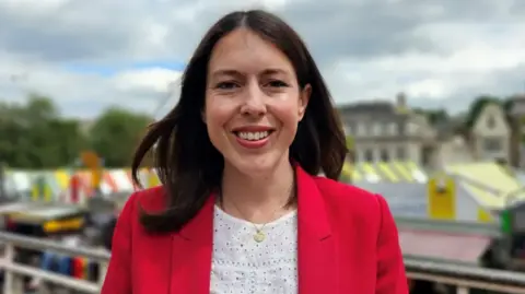 Paul Moseley/BBC Alice MacDonald smiles directly at the camera as she is photographed outside, with Norwich market visible in the background. She is wearing a red jacket and white blouse, with gold necklace. She has shoulder-length brown hair.  