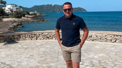 Josh Bailey A man is wearing sunglasses, a short sleeved top and shorts. He is standing on a pavement abroad with the sea behind him