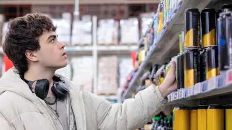 A teenage boy in a white coat with headphones around his neck looks at a shelf of yellow and black cans in a supermarket