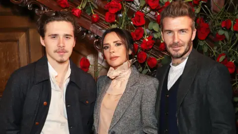 Getty Images Brooklyn Beckham, Victoria Beckham and David Beckham standing in front of wooden panelling decorated with red roses. They are wearing smart casual clothing. Victoria Beckham is wearing flesh pink blouse with ruffled collar