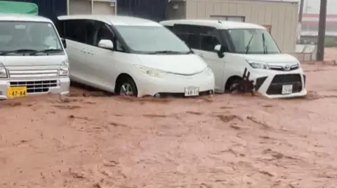 NHK Brown floodwaters flow down a road in central Japan. Three white cars are parked with water up to their wheels and bumpers.