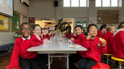 Phil Bodmer/BBC A group of primary school students, dressed in red uniforms, sit on either side of a long dining table, eating and drinking. Some smile at the camera.