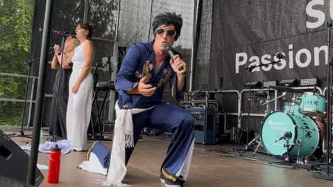 Connor Bennett/BBC A man dressed as Elvis Presley with big glasses and hair singing into the camera, with the majority of the stage behind him. Also on the stage are two women backing singers and drum kit.