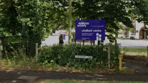 Google A blue sign with "epilepsy society" and "chalfont centre" written on it. It sits above "Chesham Lane" road sign. There is a pavement in the foreground and a large building with an archway in the background. Trees and hedges surround the signs. 