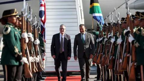 Reuters Prime Minister Sir Keir Starmer lands in South Africa and is greeted by a South African official. The men walk along a red carpet in front of white stairs leading down from a plane, past lines of people in green uniforms holding rifles.