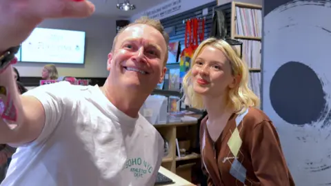A man in a white t shirt poses for a selfie with a young blonde woman in a brown cardigan