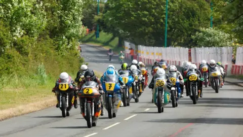 A group of motorcyclists on classic racing bikes with yellow plates in the front on a section of the Billow Circuit, which has barriers to the right and a grass bank to the left.
