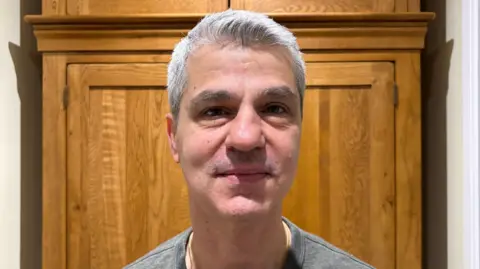 Martin Heath/BBC Andreas Kaccouris with short white hair, smiling at the camera while wearing a yellow shirt and grey sweater. There is a wooden panel behind him.