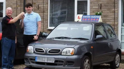 Steven Hunter, a man in his 60s who has a shaven head and wears blue jeans and a black shirt, passes his car keys to Jack Price, a man in his 20s who has brown hair and wears a light blue polo shirt and black jeans. They are standing outside a house next to a small grey car with a driving instructor "L" sign on the roof.