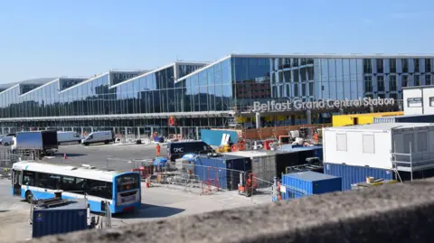 The new Belfast Grand Central Station building with parts under construction. An Ulsterbus drives along the side of a building site within the grounds of the new station.