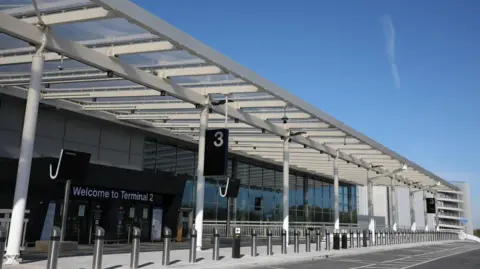 ManchesterAirport The forecourt of Terminal 2, with bollards seen running across a pedestrian area in front of parking bays. A terminal sign can be seen in front of the entrance way on a sunny day. 