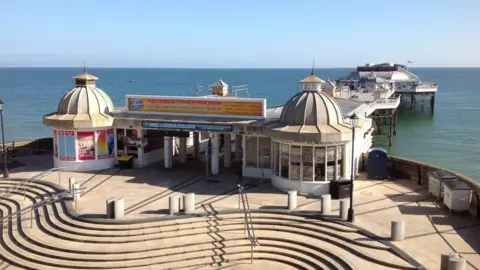 The entrance to Cromer Pier. It has steps in front of it and two small buildings either side of the entrance.