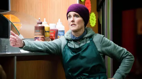 Heather Peace appearing in EastEnders, wearing a green hoody and apron and a purple beanie hat, leaning on the counter of a sandwich van.