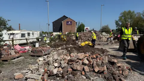 Image shows a large pile of bricks and rubble. There are around seven people in high vis jackets helping with the clean up. In the background there is an even bigger pile of dirt and rubble.
