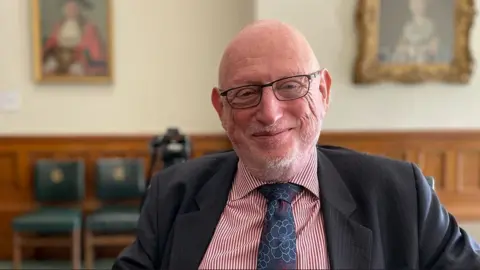 BBC Max Caller, wearing glasses and a black suit with a blue and white striped shirt and blue tie, sitting in a large room with paintings on the wall behind him.