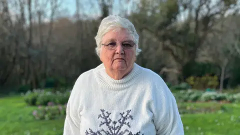 BBC A woman who looks sombre wearing a white turtle neck with a dark grey snowflake in the middle. She is standing outside with grassland and brown trees behind.