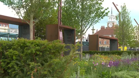 Stockton Council Three steel archways are set behind a green area planted with colourful flowers and leafy green hedges. There id a red-bricked clocktower in the back.