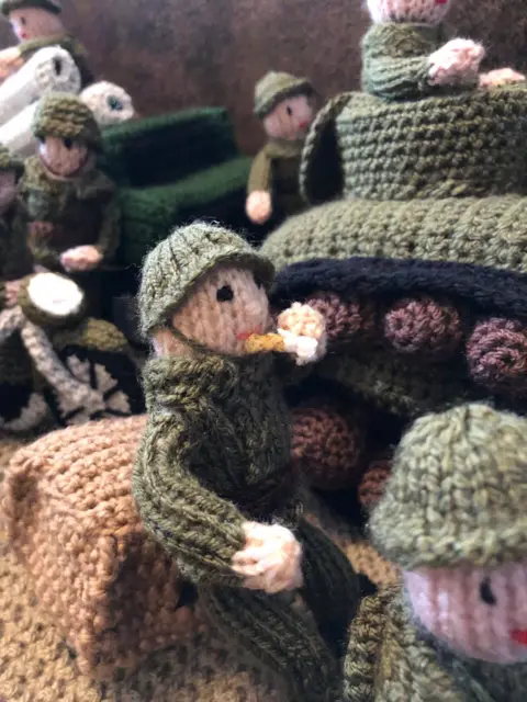 The Longest Yarn A knitted solider is sitting next to a tank smoking a cigarette. Behind him are more soldiers wearing green outfits and helmets.