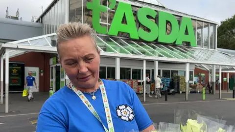 Asda A woman in a blue T shirt with a white rose stands outside a supermarket with a sign that reads t'Asda