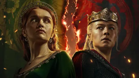 HBO Twee personages uit Dragon House met vuur op de achtergrond, een vrouw met rood haar in een groene jurk, de andere vrouw met blond haar in een kettingjurk en kroon