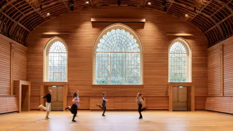 A wooden building with large domed windows. The building has people running across it, kicking their legs up behind them.