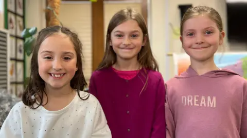Molly, Angelina, Ruby are standing indoors in front of a classroom display. They are wearing casual clothing in shades of white, pink, and maroon. Behind them, there is a wall with educational materials and a large map partially visible on the right side.