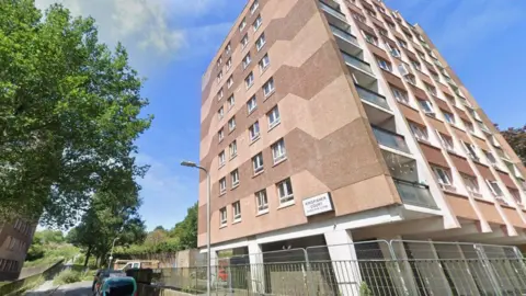 A tower block of flats, with tree next to it. 