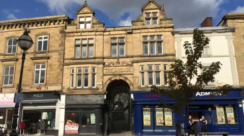 The Arcade The front of Dewsbury Arcade. The stone facade features a series of long mullioned windows over three storeys and a central arched entrance on the ground floor. High street shops and businesses line the street on either side of the entrance.