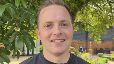 John Fairhall/BBC Tyler Whitnall smiles at the camera. He has short light hair and wears a dark T-shirt.