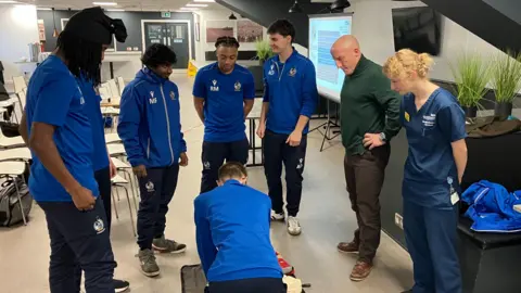 A boy is knelt down next to a medical training dummy, leaning over it performing CPR. Around him there are four other students watching, a man in a green jumper and a woman who is wearing an NHS uniform. The students are wearing blue tops and trousers with Bristol Rovers logos on them. 