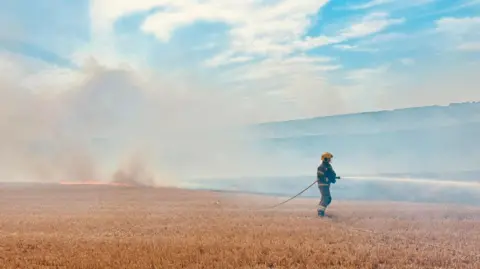 DWFRS A firefighter stands in the stubble of a harvested crop field directing a hose towards the right of the picture. Behind him to the left is another fire and smoke fills the air.