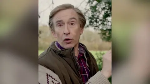 A man, Alan Partridge, wears a checked shirt and a jacket with patterned sleeves. He is standing outside.