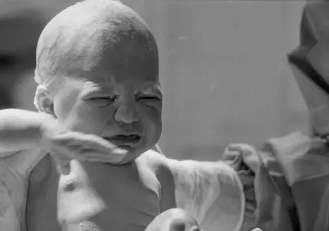 Francis Sanders Ester moments after being born. The black and white photograph shows her being held by a medic. She is crying.