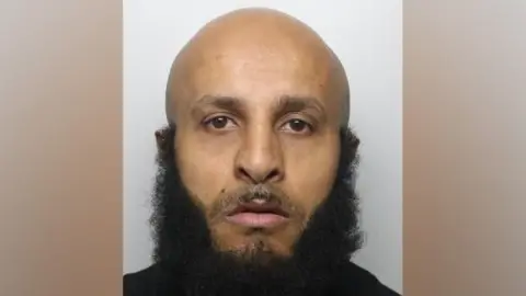 A mugshot of Waleed Ali who is bald and has a thick, black beard along his jaw. There is also a bit of facial hair above his lips.