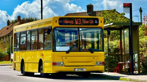 A yellow bus, with writing at the top, by a bus stop, with a row of houses behind. There is also foliage around the bus stop. 