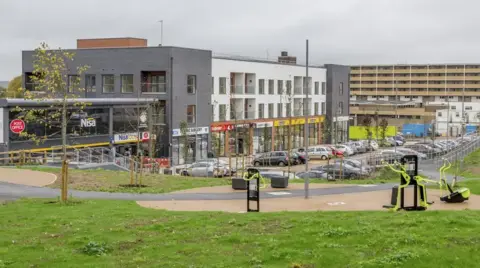 ENGIE A row of shops with flats above them. It is a new building. There is a playground on a grassy area in the foreground of the picture.