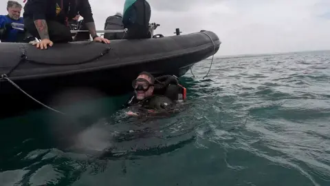 Dr Raymond Ward A man in a diving suit smiles as he bobs in the sea by a power boat.