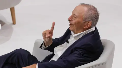 EPA Larry Summers, sentado de perfil en un sillón gris, vestido con traje azul marino y short blanco, sonríe mientras levanta el dedo índice de su mano derecha.