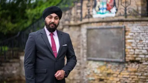 Mr Sohal is wearing a suit with a pink short and darker pink tie. He wears a turban and is stood outside by a brick wall.