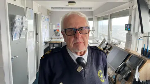 A man with short white hair and rectangular glasses is wearing a v-neck jumper with a yellow logo, a white shirt and black tie. He is pulling a serious expression and an office room can be seen behind him. Outside the window you can see the sea.