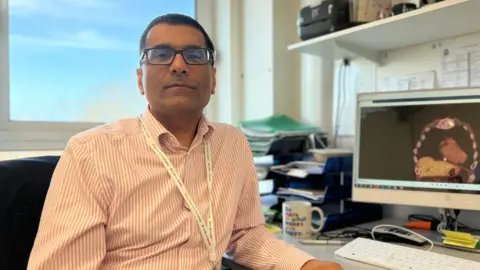 Rubin looks at the camera as he sits next to his work desk where a computer showing scan. He is wearing a pink striped shirt and has a rectangular glasses on. Rubin has short black hair.