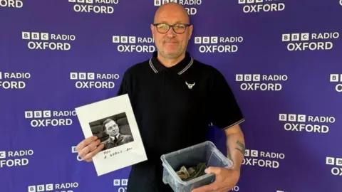Danny Jones is wearing a black polo shirt and is bald. He is holding an image of Sylwester Godlewski in one hand, and parts of the spitfire in the other. Behind him is a purple wall, with 'BBC Radio Oxford' written in white on it.