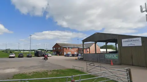 Google A gate leading to a car park with open barn-type building and a red brick club house, with rugby pitches in the distance. A sign says 'Welcome to Chinnor RFC'
